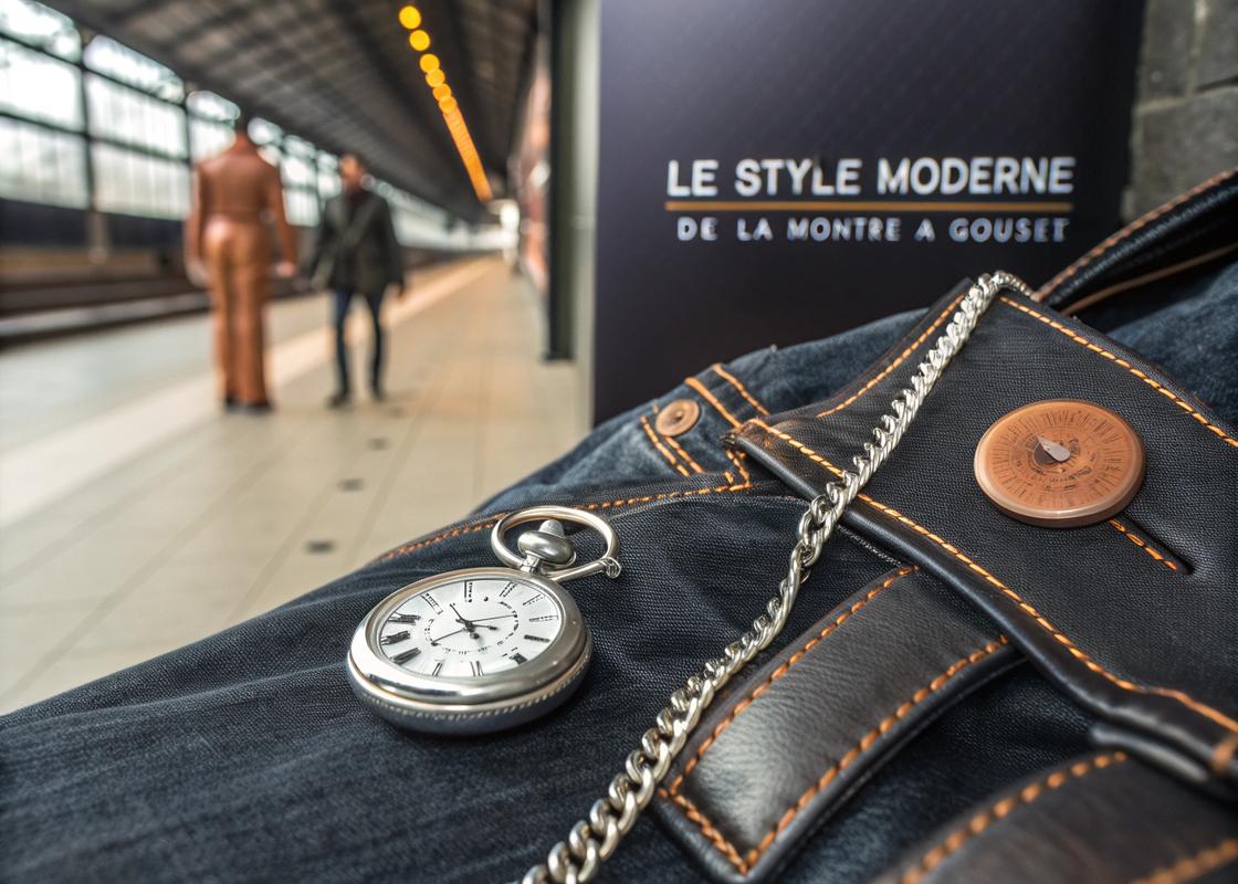 Montre à gousset en argent mat accrochée à une boucle de ceinture sur un jean, avec des éléments steampunk en cuivre et une veste en cuir en arrière-plan.